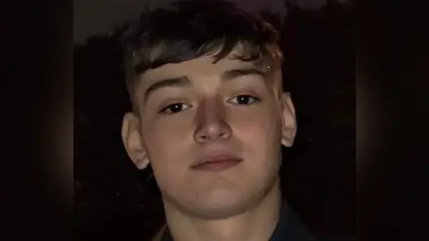 A man with dark brown hair and eyes looks at the camera, in front of a dark background