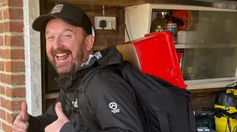 A laughing Stu Skinner gives a double thumbs up to the camera. On his back is a backpack with a red rectangular post box poking out the top. He has a full brown beard and is standing side-on to the camera. He appears to be in a domestic garage and is wearing black hiking gear and a black baseball cap. 
