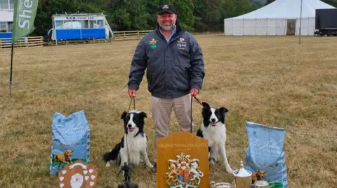 Ben is standing in a field holding the leads of his two black and white sheepdogs who are either side of him. There are two wooden shields in front of them and two bags of blue dog food on either side of the dog.
