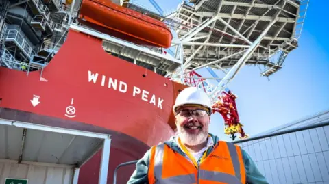 Dale Baxter/BBC A smiling man stands on a ship, wearing glasses, a hard helmet and a hi-vis. Behind him rising a red superstructure, with the words WIND PEAK written on it in white letters.