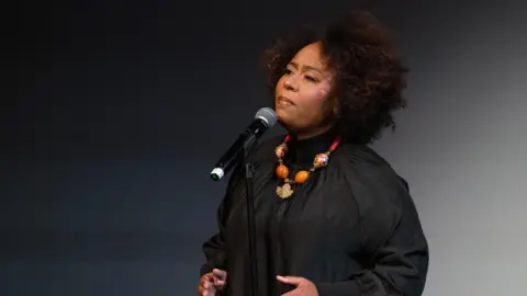 Emily Mudie Photography A woman in a black top sings on stage. She wears a colourful necklace. There is a microphone in front of her.