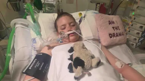 Felicity-Jo Rowlett-Howes with her eyes closed in a hospital bed with many wires attached