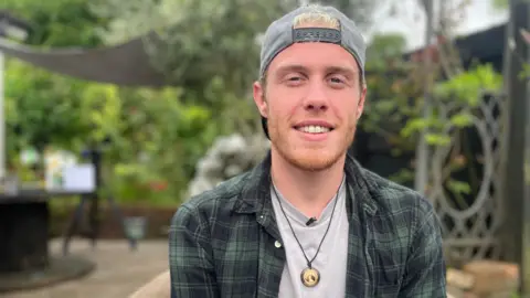 A man with blonde hair and a ginger beard wearing a grey backwards baseball cap, a grey t shirt, green check shirt and a necklace. He is smiling and looking at the camera, and behind him is a garden with green foliage.