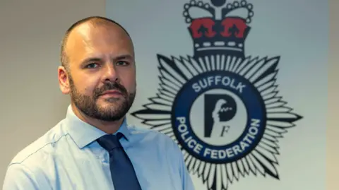Suffolk Police Federation A head and shoulders image of Matt Paisley. He is wearing a light blue shirt and a navy tie. He is standing in front of a large Suffolk Police Federation emblem, which is on the white wall behind him. He is looking into the camera. 