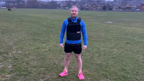 Pete Grimwade Pete Grimwade in his marathon running gear of blue, longsleeved top, black shorts and pink trainers, wearing his 10kg pack strapped to his chest - standing in a field 