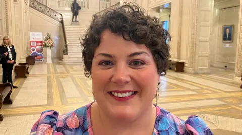 Maria Thomasson is standing in the middle of the Great Hall at Stormont. She is a young woman, with short, brown curly hair and she is wearing red lipstick and a pink and blue floral pattern dress. She is smiling at the camera.