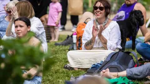 James Mulkeen A group of people - the audience of a festival - seated on grass. At the centre is a smiling woman with long dark hair, wearing sunglasses, a white blouse and long white skirt covering her crossed legs. Her arms are tattooed and she is clapping her hands. Behind her is a black dog. 