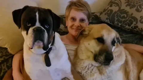 Caroline Dewson, a woman with blonde hair, sitting on a sofa with her arms around two large dogs. One of the dog is sandy-coloured, the other is brown and white and looks like a boxer-cross.