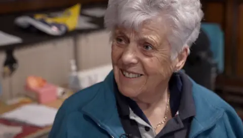 A woman, who is wearing a navy polo shirt and teal jumper, is smiling at the interviewer in this still from the interview footage.