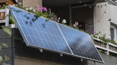 A solar panel hanging on a balcony, which also contains plants and flowers.
  