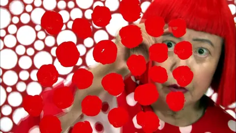 Yayoi Kusama seen looking through a clear screen with red spots painted on it, with red and white clothes and background