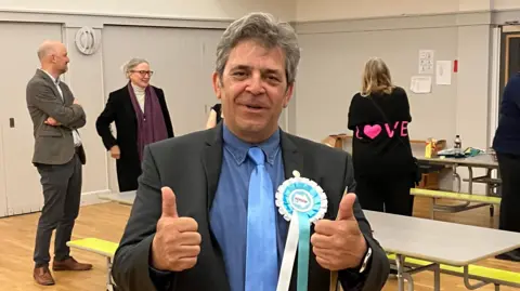 Lyndon has dark grey wavy hair and wears a dark grey suit, light blue shirt, and a teal tie with a Reform UK rosette on his left lapel. He has both of this thumbs up celebrating with other people behind him in the counting hall. 