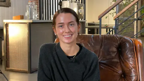 Betsy is sat in a large leather chair in a salon. Behind her is a counter with a large water jug on it, and a set of stairs. She has brown hair and wears and dark grey jumper. She is smiling at the camera. 