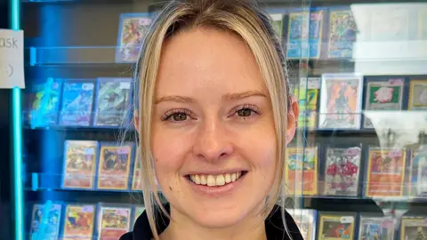 A young blonde woman is smiling, behind her is a wall covered in Pokemon cards behind a class case.