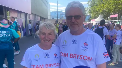 The photo shows a women and a man looking and smiling at the camera. The women is on the right and the man is on the left. The man has his arm around the women's waist. They have white t-shirts on with Team Bryson written on the front with the Team GB branding. They are stood in front of a small crowd of people on a small street setting. 