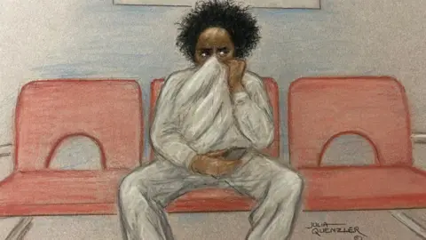 Julia Quenzler / BBC A court sketch of Axel Rudakubana who is depicted wearing a grey tracksuit and sitting on a red chair. He has black hair which is sticking up and he is holding the top of the sweatshirt over his mouth and nose while looking to the right.