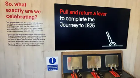 This is an interior photograph of one of the carriages on the train - there are levers at the bottom of the picture. Above them is a screen that that says: "Pull and return a lever to complete the journey to 1825."
There is aso an explanation next to the screen of how 200 years ago saw the opening of Stockton & Darlington Railway in 1825.