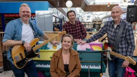 A four-piece band with an upright piano including a man holding a wooden bass guitar and another with an electric guitar