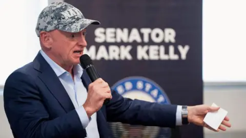 Reuters Senator Mark Kelly wearing a baseball cap and a suit in front of a United States military emblem