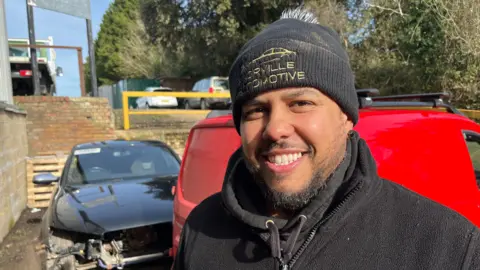 John Devine/BBC David Norville, has a black woolly hat on his head and black fleece, he has a wispy dark beard and moustache and is smiling, behind him are cars, one with damage.