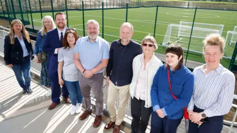 A group of adults stand in a line in front of a green sports pitch which has white football nets on it. They are all smiling on a sunny day.
