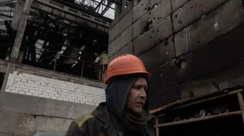 A workman in an orange safety helmet walks through a large charred industrial plant.