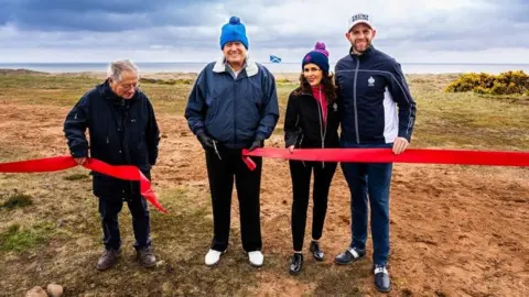 Trump International Trump cuts a red ribbon alongside two men and a woman wearing wolly hats and warm clothing againsta stormy sky.