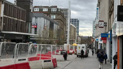 Newcastle's Northumberland Street. The road is one of the city's main shopping street with high street shops and lots of people milling about. There are large red and white bollards with wire fencing that is blocking off the central section of the street.