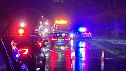 Emergency services vehicles with flashing lights, police tape, and a queue of cars.