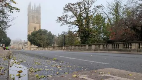 Lucie Johnson Looking along the High Street from the bridge, a large tower on the right hand side of the road. Leaves and puddles are on the road.