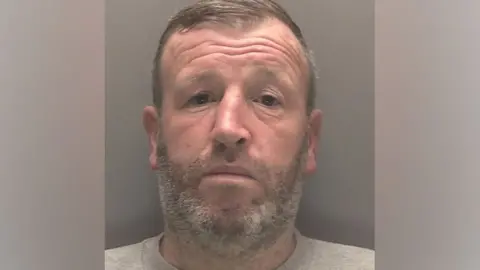 Merseyside Police Bearded Brian Spencer looks ahead, unsmiling, in his police mugshot. He is wearing a grey top.