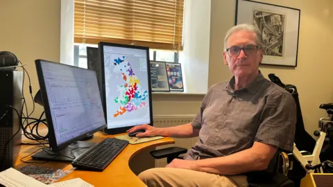 A man in his sixties wearing reading glasses, a brown short-sleeved shirt and lighter brown trousers sits behind his desk with several computer screens in front of him. One on the right displays a map of England and Wales with coloured dots on it. The screen of the left computer is just visible and shows a couple of graphs. In the background is a window, radiator, framed awards and a bike machine. 