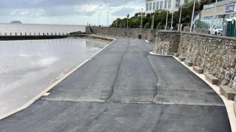A dark grey, tarmac-like surface runs along the side of the marine lake, with a white edge and a stone wall on the other side. You can see the sea beyond the edge of the lake