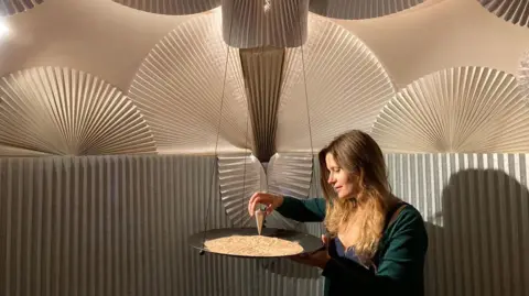 Discovering 42 Large fans span out across the room. A woman stands in the middle holding a metal pendulum that is drawing in sand on a large suspended dish.