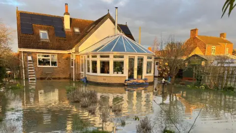 Supplied Water submerges the garden of a home. Some long strands of grass and bushes stick out of the water. Sandbags are piled up at the entrance to the house's conservatory. Fire rescue crews can be seen in walking through the water.
