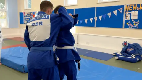 Two men wearing blue judo outfits grapple with one behind the other ones back. Two blue mats are in front of them.