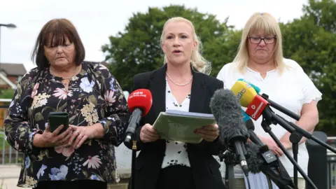 PA Media Three women, one with shoulder length brown hair and a fringe, another in a black suit with long blonde hair tied up, and a third with long blonde hair with a fringe, standing beside each other in front of media microphones. 