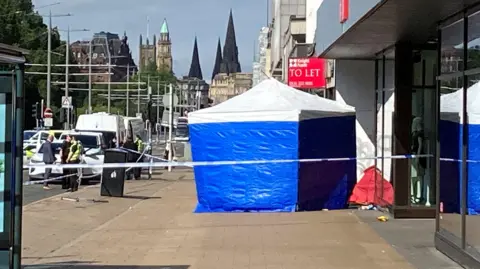 A blue and white police tent behind a corden on a pavement with police officers standing next to a police car