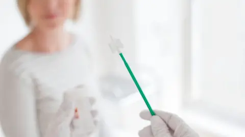 A doctor or nurse's hand can be seen holding cervical screening equipment, while an out of focus woman sits nearby