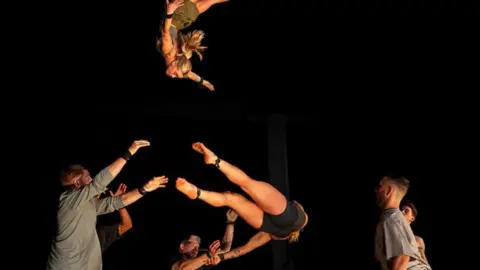The 10,000 Hours acrobat troupe. A person is flying through the air towards performers on the ground who are swinging another person around