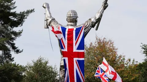 A tall statue with a large union jack attached to its back. There are several smaller union jacks and St George's crosses around it.