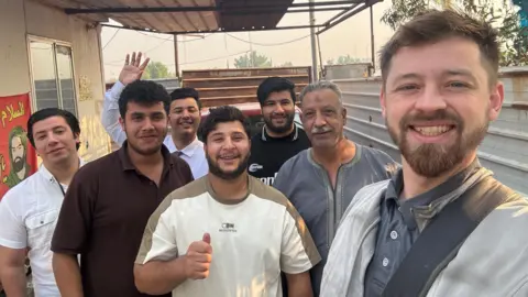 We Hate The Cold Harry Jackson, to the right of the picture, stands by a building in Iraq with six people. They are all smiling and looking at the camera. 