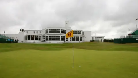 PA Media A golf course, in the foreground is the green, flag and a golf ball near the hole. In the background is a large white building. On the edges of the image are the start of stands on either side of the course.