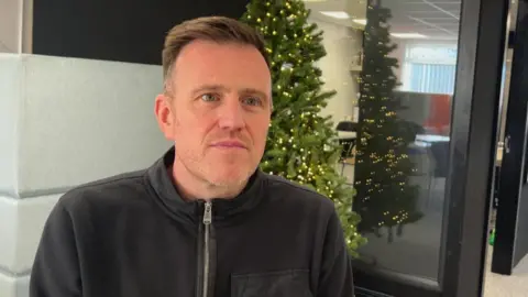 A man with short brown hair and black zip-top with black framed interior doors behind him and a green christmas tree with lights on