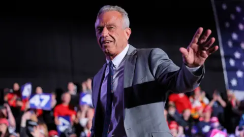 Reuters RFK Jr, que tiene cabello gris, viste un traje gris, con camisa blanca y corbata estampada azul marino, mientras saluda a la multitud en un mitin de Trump en Michigan.