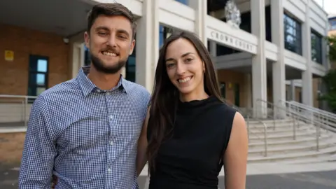 Ben, a man with brown hair and a short beard wearing a shirt, and Emma, a woman with long dark hair and wearing a black sleeveless top, outside court