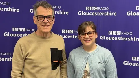 A dad and daughter stand next to each other in front of a BBC Radio Gloucestershire banner. They both wear glasses and smile looking at the camera. The girl has a fringe and wears a light blue cardigan while the dad wears a beige jumper. 