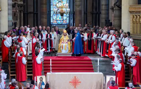 Gareth Fuller / PA Media A wide view shows Mullally sitting in the middle of the congregation with red and white gowned clergypeople all around her.