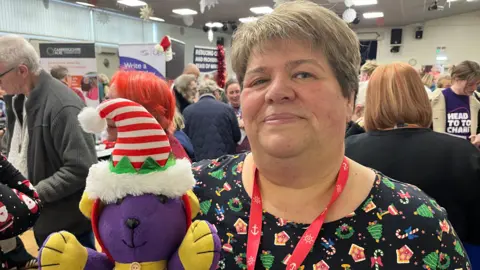 John Devine/BBC Sam Clark has short reddish hair and is wearing a Christmas top with various festive symbols on it. She is holding a soft toy purple dog which has yellow paws and a yellow collar and which is wearing an elf hat. Behind her are lots of people in a function room with information signs for various trade stands.