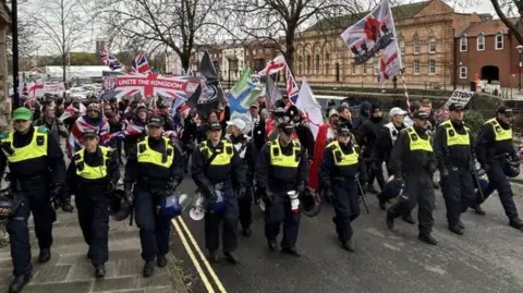 A line of police in neon vests in front of a crowd of people holding different variations of the Union Jack, including some with slogans like "Unite the Kingdom" and . A row of red brick buildings can be seen in the background.